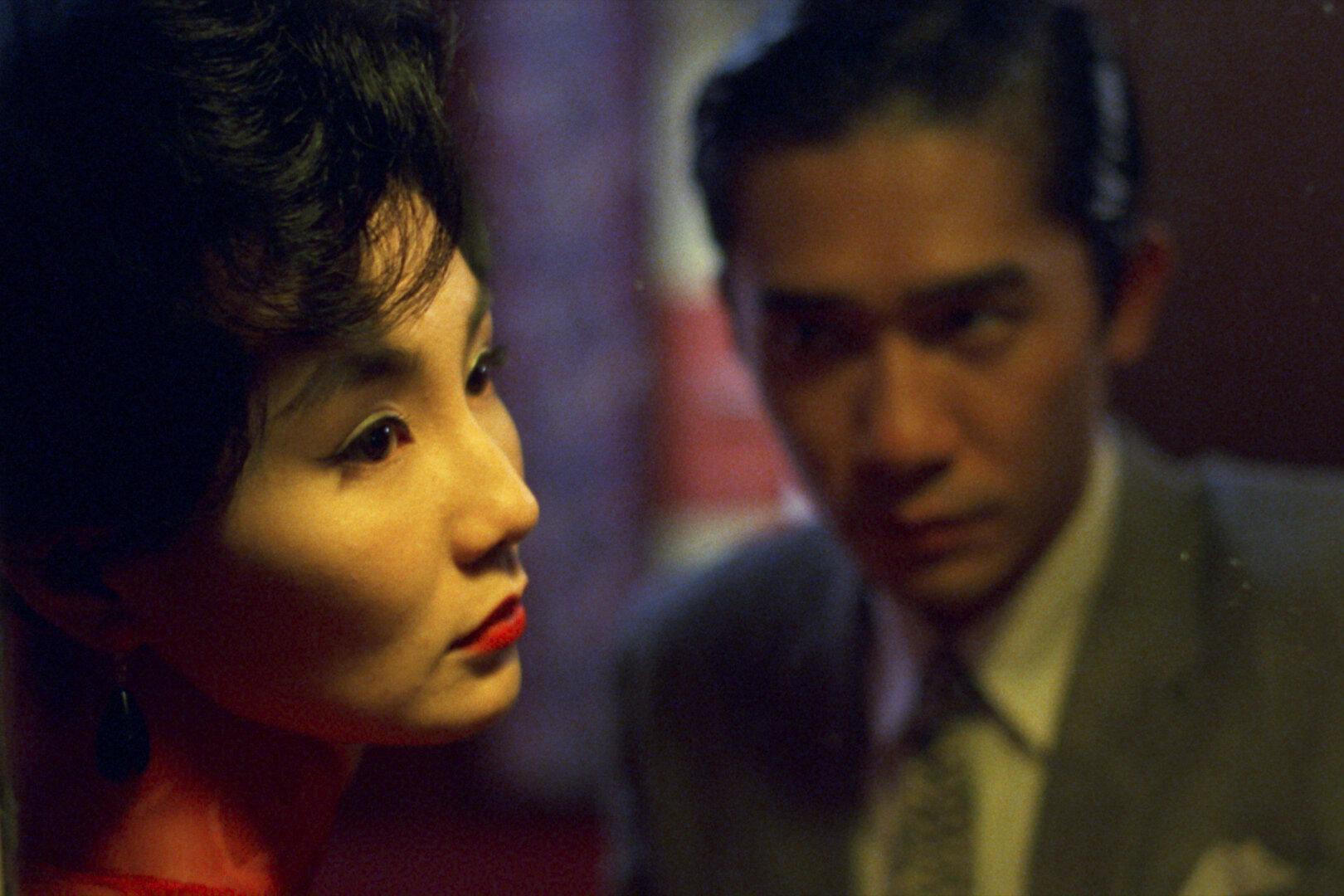 A woman (Maggie Cheung) is framed in close-up with an out of focus man (Tony Leung) gazing at her from the right side of the frame.